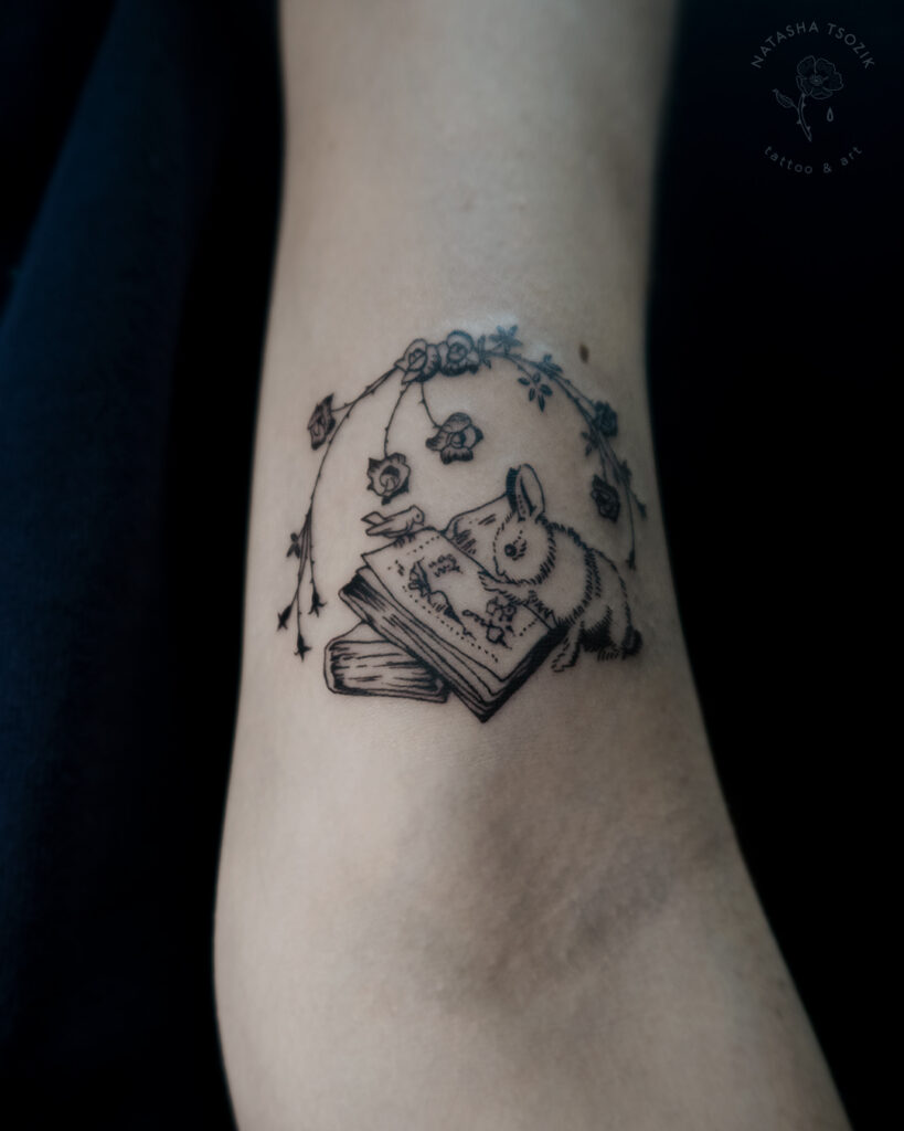 A small black&white tattoo on an inner bicep. A rabbit reading a book, surrounded with roses.